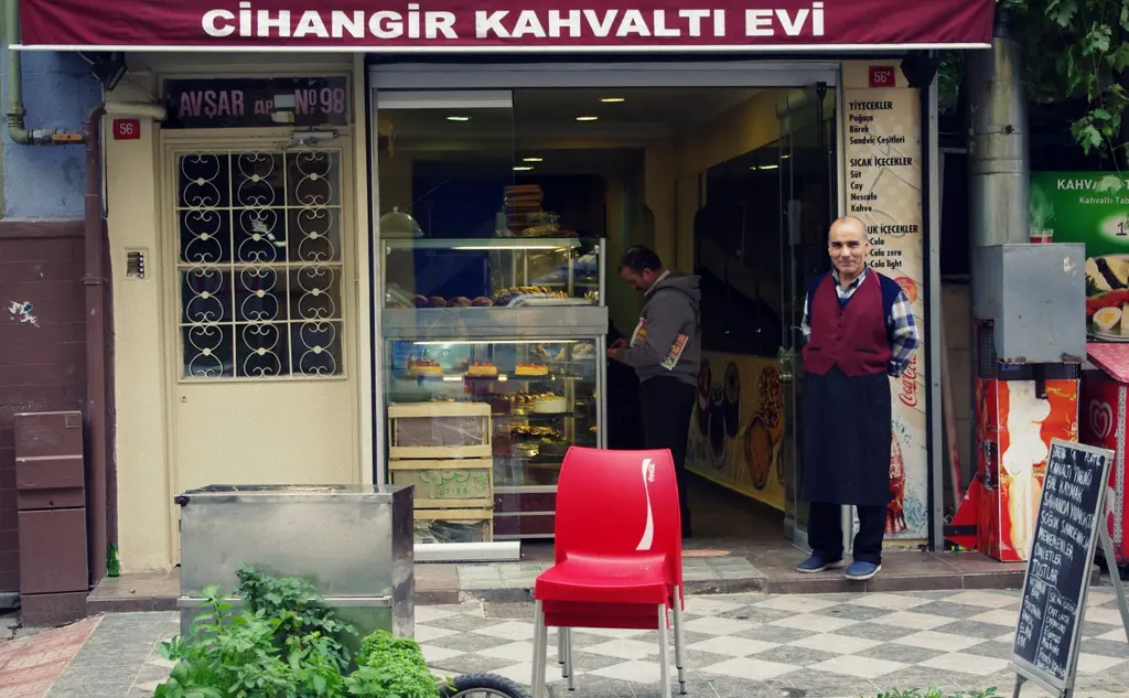 Cihangir'de bir kahvaltıcının önü; kırmızı tabela, vitrinde yiyecekler, kapıda önlüklü bir adam ve kaldırımda kırmızı sandalye.