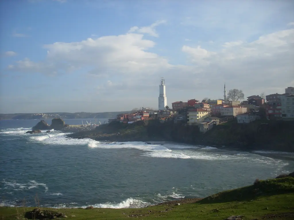 Rumelifeneri'nde deniz feneri; tepedeki köy evleri arasında beyaz kule, kayalık kıyı, dalgalı Karadeniz ve bulutlu gökyüzü.