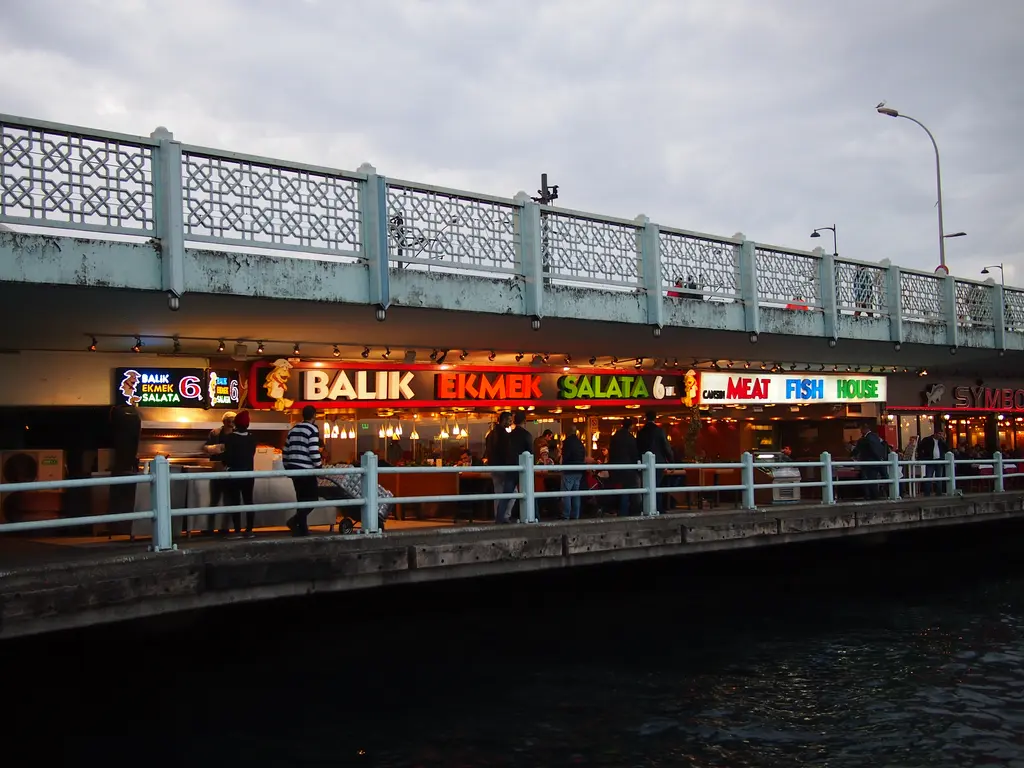 Galata Köprüsü'nün alt katı akşamüstü; Balık Ekmek, Salata ve Fish House yazılı ışıklı tabelalar ve üst katta korkuluk.