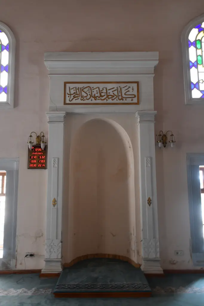 Şah Sultan Camii'nin mihrabı; beyaz niş, üstünde yazılı levha, iki yanında renkli camlı pencereler ve mavi halı.