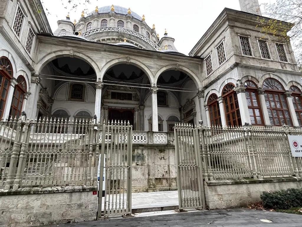 Nusretiye Camii'nin girişi; kemerli revak, iki katlı yan bina, beyaz demir parmaklıklar ve üstte kubbe.