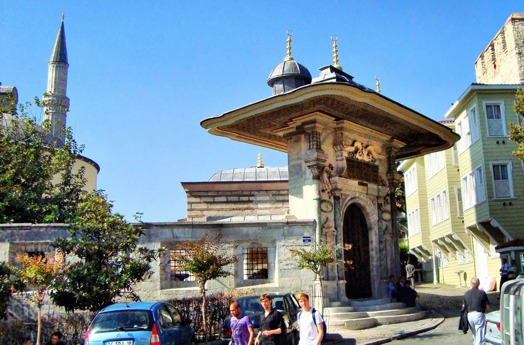 Soğukçeşme Sokağı'nın başında III. Ahmed Çeşmesi; geniş saçaklı mermer yapı, solda minare, sağda ahşap evler, önde insanlar ve arabalar.