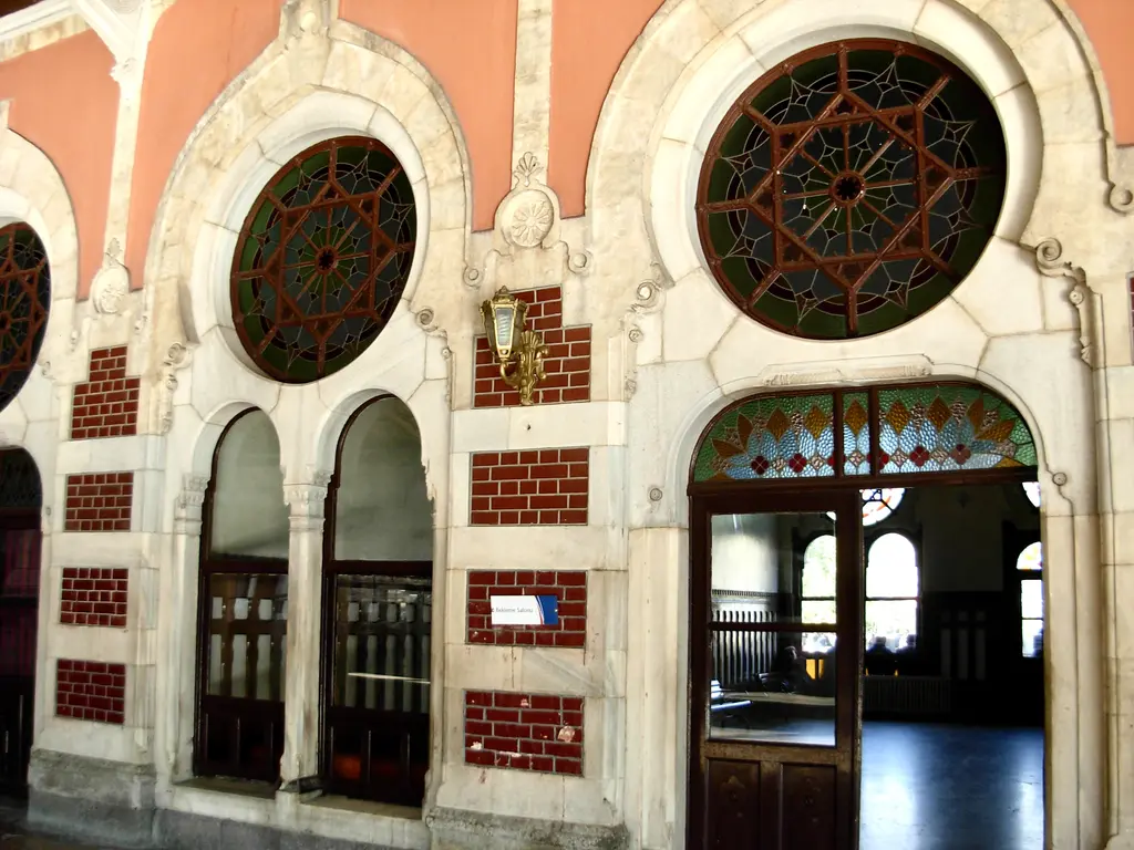 Sirkeci Garı'nda kiremit turuncusu duvar; krem taş çerçeveli iki gül pencere, aralarında renkli camlı sivri kemerin altında koyu ahşap kapı ve duvarda küçük fenerler.