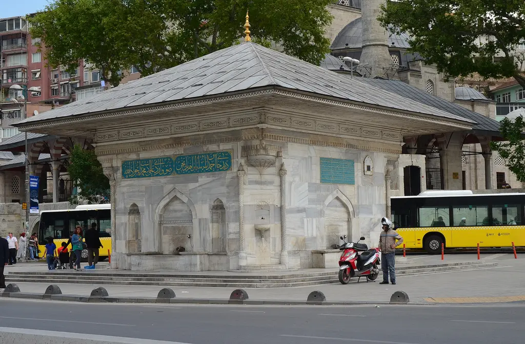 Üsküdar III. Ahmed Çeşmesi; mermer gövde, yeşil kitabe levhaları, arkada cami kubbesi ve sarı otobüsler.