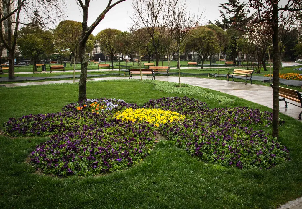 Göztepe Parkı'nda çiçek biçimli tarh; mor ve sarı hercaimenekşeler, çimenlik, banklar ve arkada yürüyüş yolu.