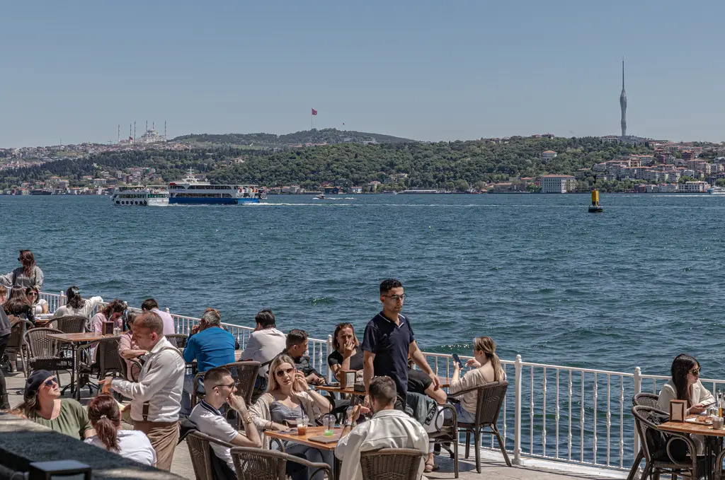 Dolmabahçe kıyısında terasta oturan insanlar; önde masalar ve korkuluk, Boğaz, karşı kıyı ve ufukta kule.