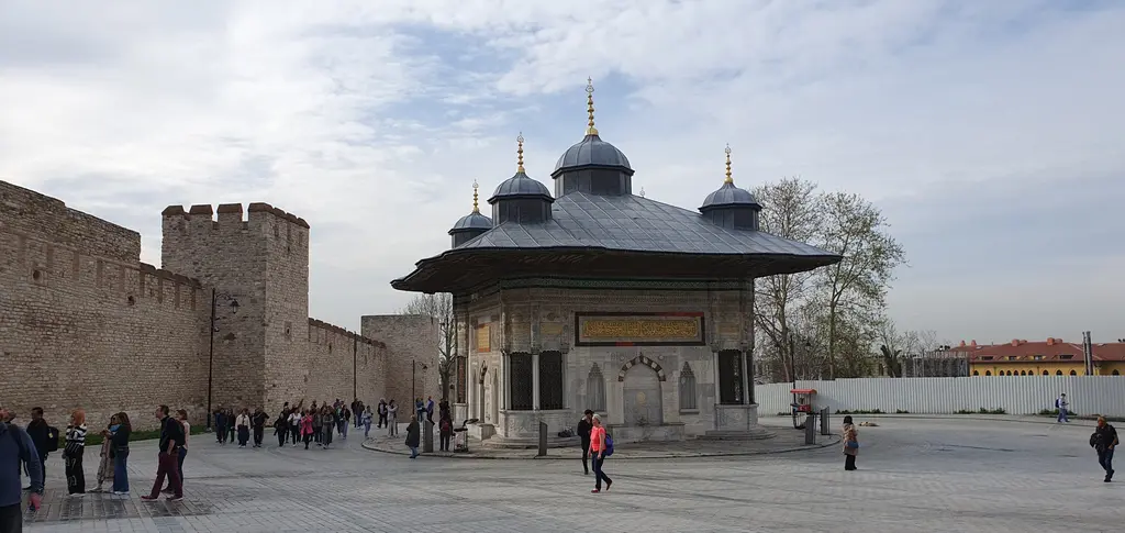 Geniş açıyla çeşme ve meydan; solda saray duvarı, ortada çeşmenin dört cephesinden ikisi, sağda ziyaretçiler ve ağaçlar.