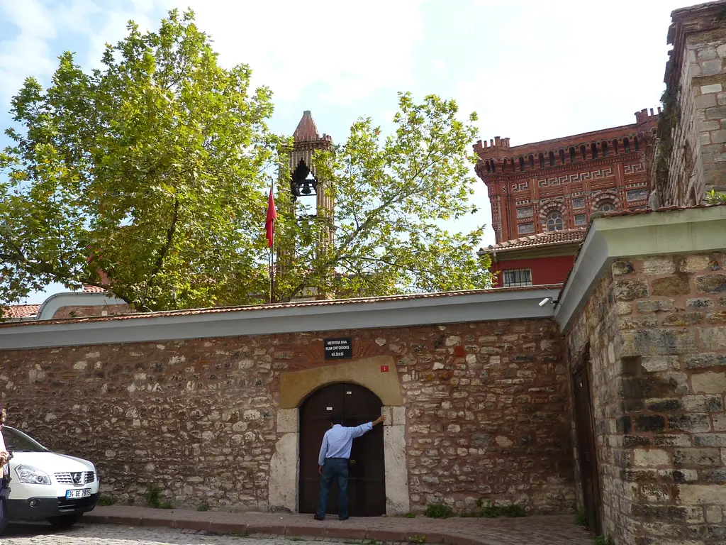Kanlı Kilise'nin giriş kapısı; taş duvardaki ahşap kapıda bekleyen bir adam, üstünde levha, arkada çan kulesi, bayrak ve Rum Lisesi.