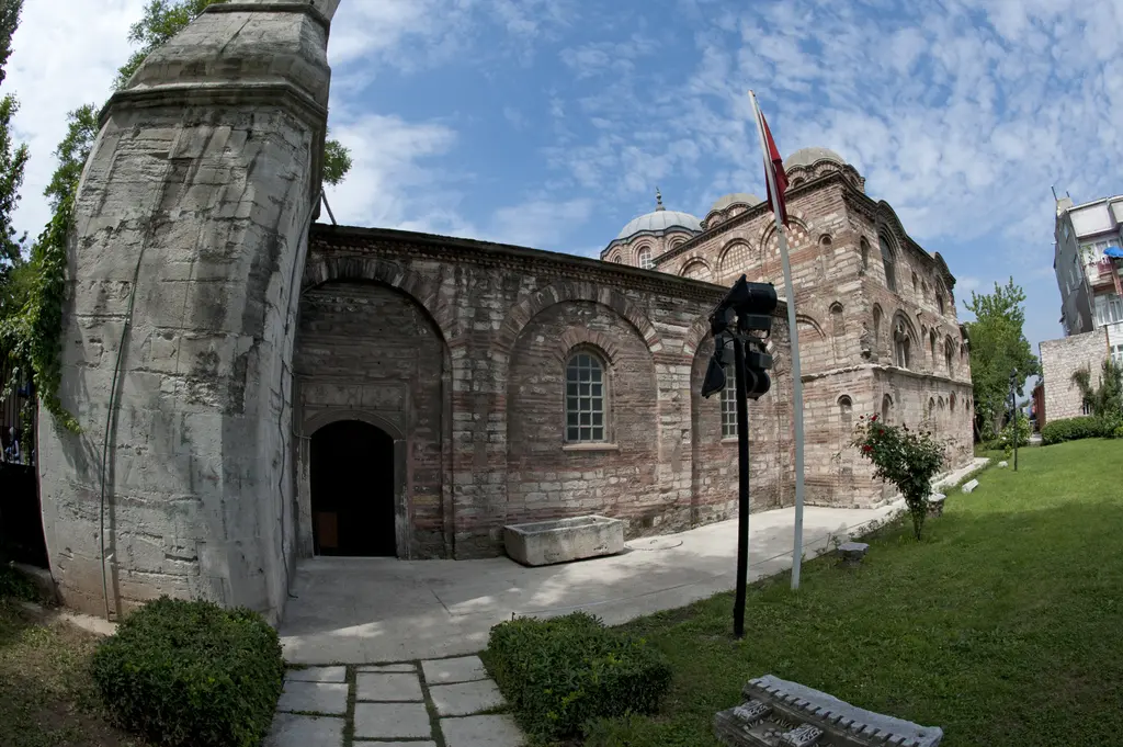 Fethiye Camii'nin avlusu; taş döşeli patika, çimenlik, minarenin yivli taş kaidesi, arkada yapının kuzey cephesi ve bayrak direği.