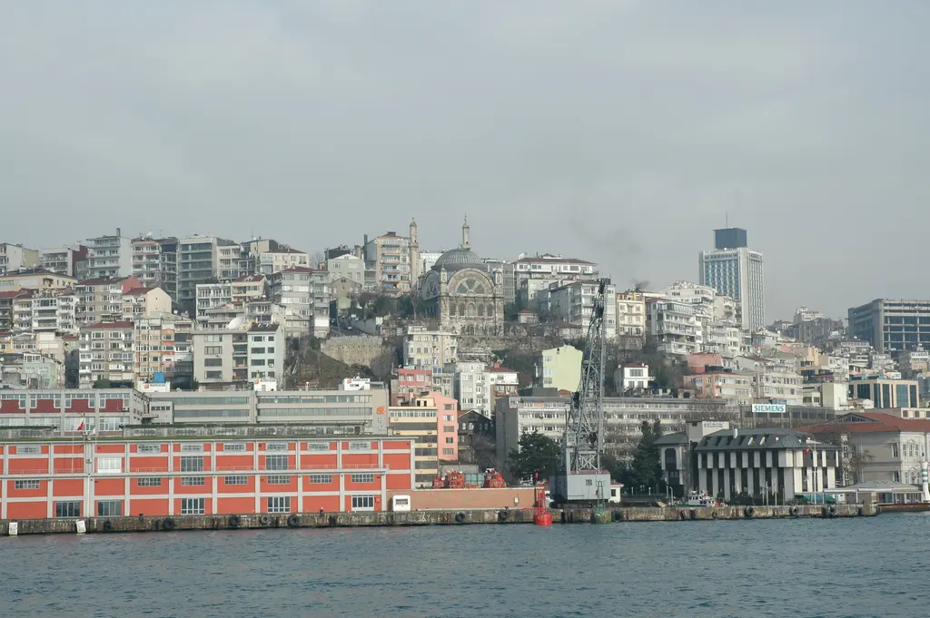 Boğaz'dan Cihangir; kıyıda kırmızı liman binası ve vinç, arkada yamaca yayılan apartmanlar ve kubbeli cami.