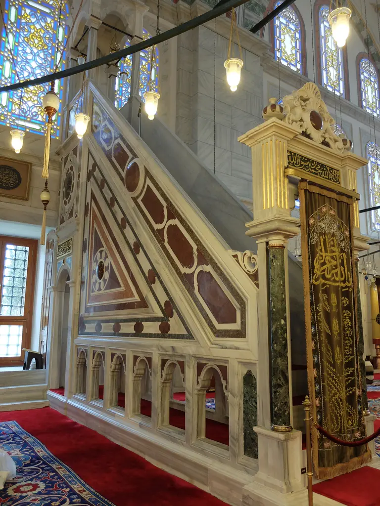 Fatih Camii'nin mermer minberi yandan; beyaz mermer merdiven, aynalıkta geometrik çerçeveler içinde koyu kırmızı porfir panolar, tepede sütunlu köşk ve önde desenli seccade.