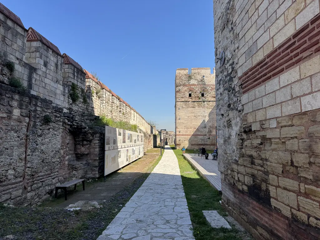 İç sur ile dış sur arasındaki koridor; taş döşeli yürüyüş yolu, kenarda bilgi panoları, bank, çim şerit ve iki yanda yükselen duvarlar.