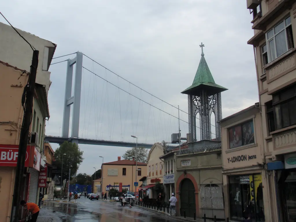 Ortaköy'de yağmurlu bir cadde; ıslak kaldırım, yeşil külahlı çan kulesi, dükkânlar ve arkada asma köprünün ayağı.