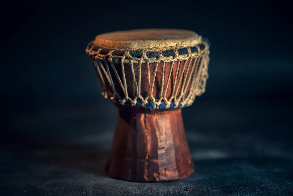Koyu bir zemin önünde ip gerdirmeli, deri derili küçük ahşap bir Afrika davulu (djembe); sıcak ışıkla vurgulanmış oymalı gövdesi.