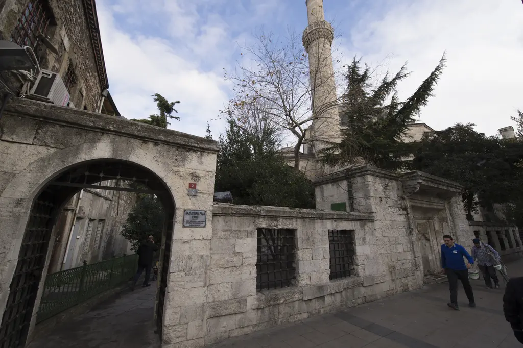 Gazi Atik Ali Paşa Camii'nin hazire duvarı; taş kemerli kapı, demir parmaklıklı pencereler, arkada ağaçlar ve minare, önde yayalar.