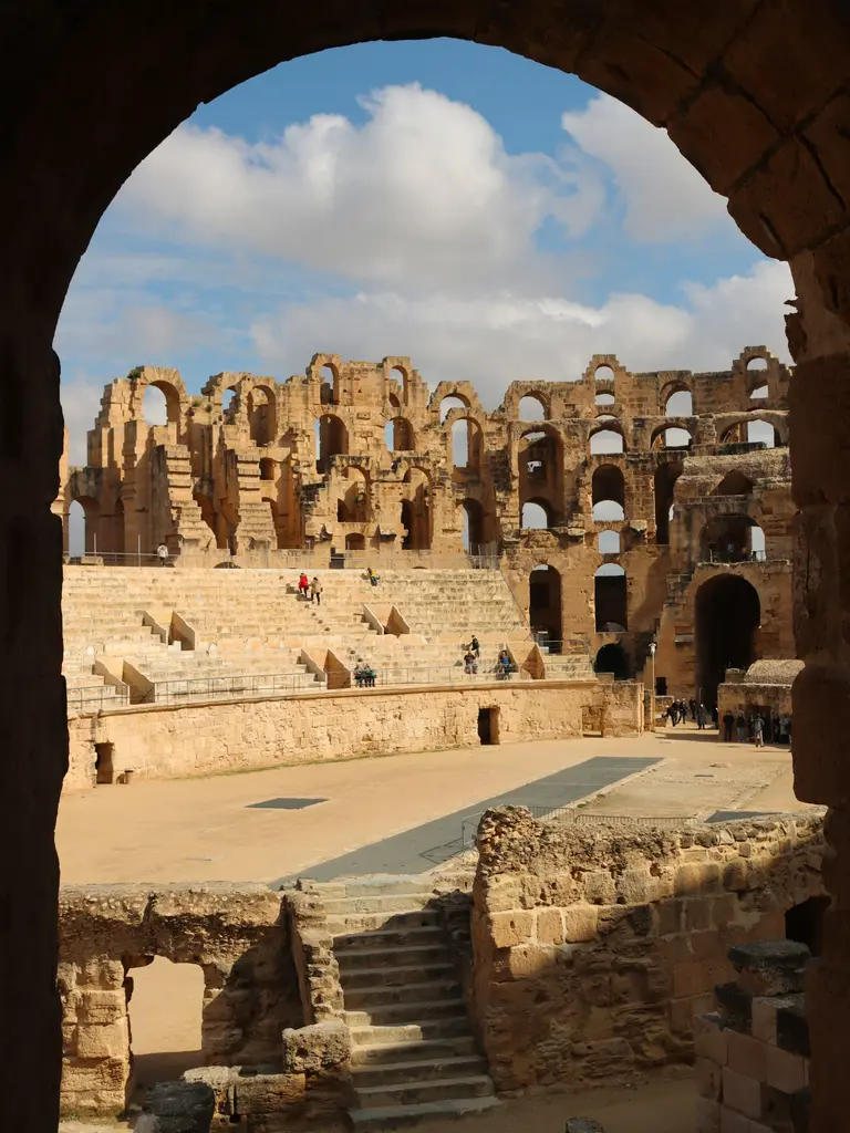 Tunus'ta El Cem Roma amfitiyatrosu; taş kemerden çerçevelenmiş çok katlı arena tribünleri ve beyaz bulutlu mavi gökyüzü.