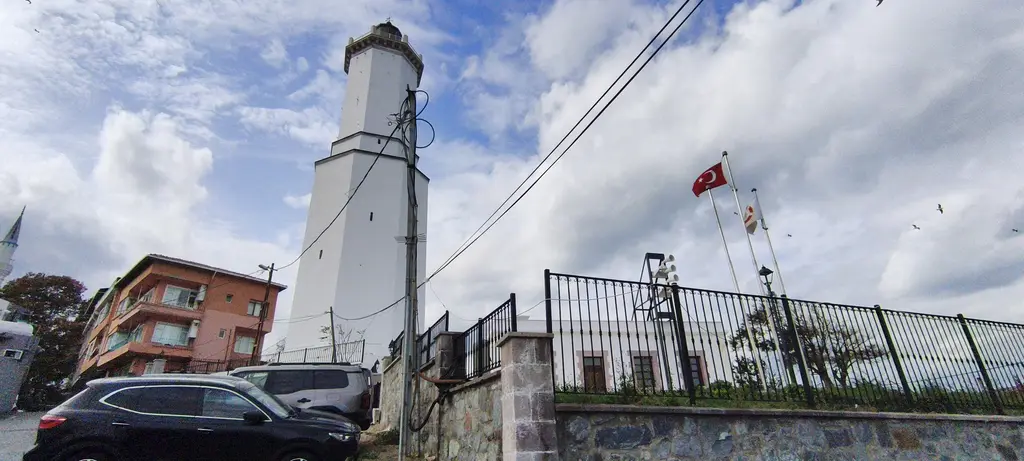 Rumelifeneri'nde fener kulesi; beyaz sekizgen kule, bahçe çiti, önünde park etmiş araçlar ve parçalı bulutlu gökyüzü.