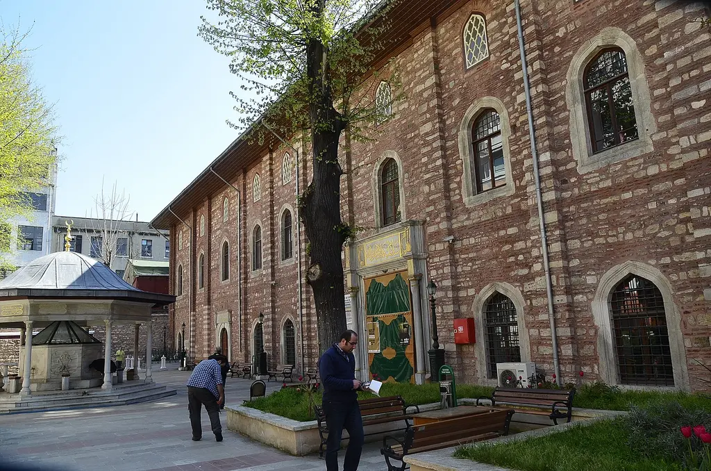 Arap Camii'nin avlusu; solda tuğla cephe ve kapı, sağda kubbeli şadırvan, ortada taş döşeme ve yürüyen insanlar.