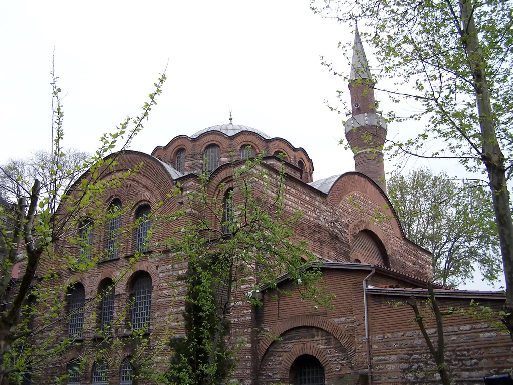 Kalenderhane Camii'ne aşağıdan bakış; kademeli yükselen tuğla kollar, dilimli kasnaklı kubbe, sağda minare ve önde taze yapraklı dallar.