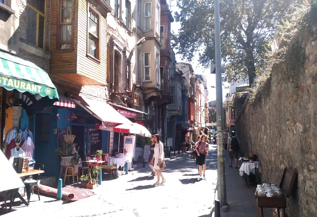 Balat'ta kafeler sokağı; tenteli dükkânlar, kaldırımdaki masalar, sağda yüksek taş duvar ve yürüyen insanlar.