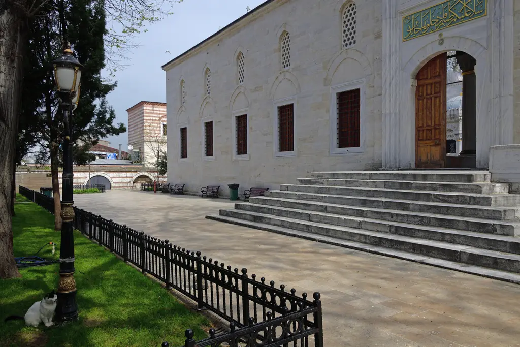 Yeni Valide Camii'nin batı cephesi; taş duvar, sıra pencereler, geniş merdiven ve demir parmaklıklı bahçe.