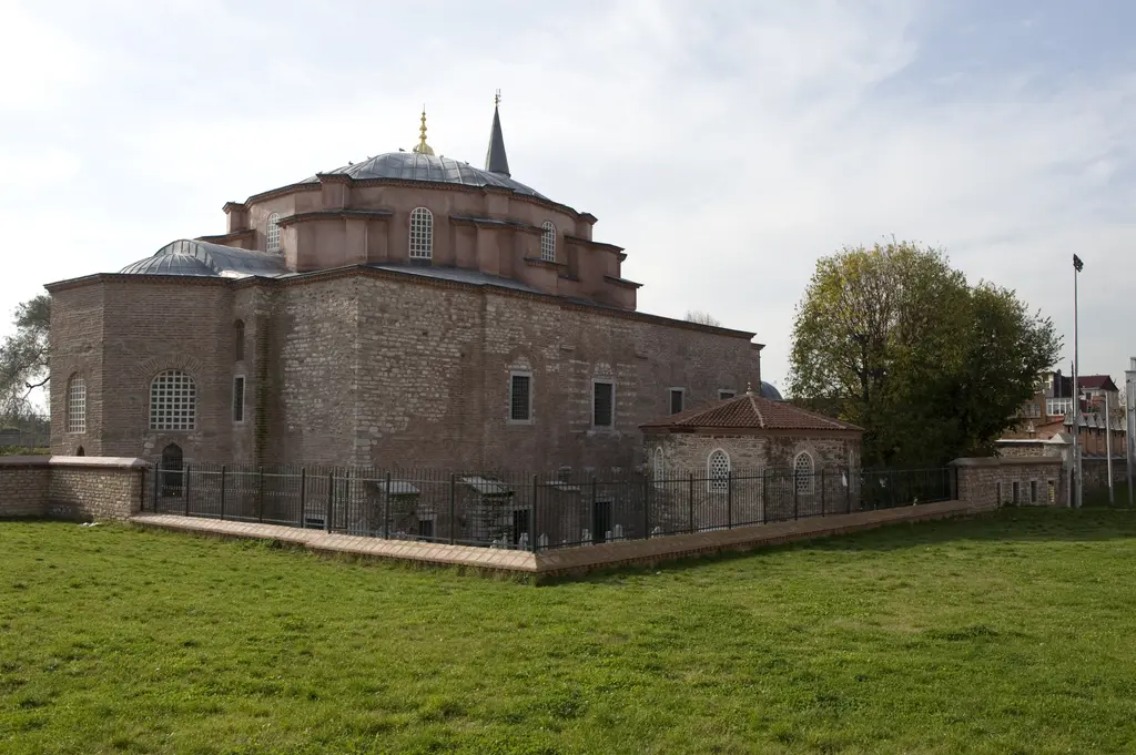 Küçük Ayasofya Camii'nin kuzey cephesi; kırmızı tuğla ile beyaz taşın sıra sıra dizildiği duvarlar, alçak kurşun kubbe, sağda minare külahı, önde geniş çim ve sonbahar ağacı.