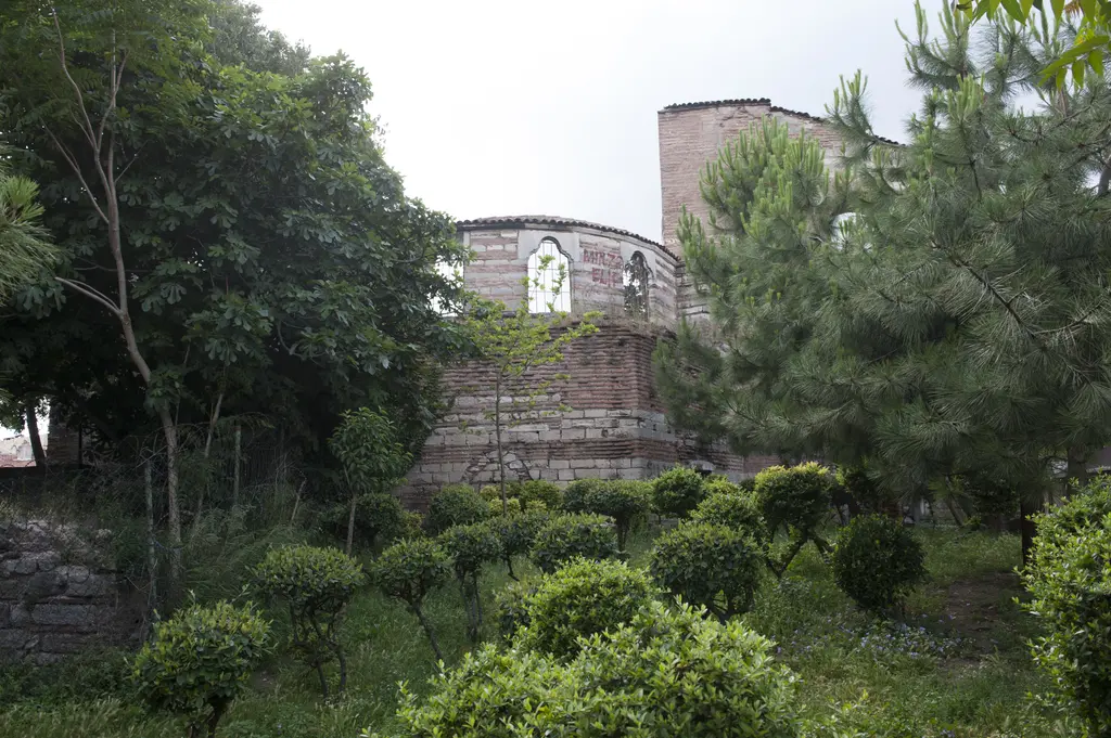 İmrahor İlyas Bey Camii bahçeden; ağaçların arasından görünen kemerli pencereli yıkık duvarlar, sağda yüksek kütle, önde çimen ve sık yeşillik.