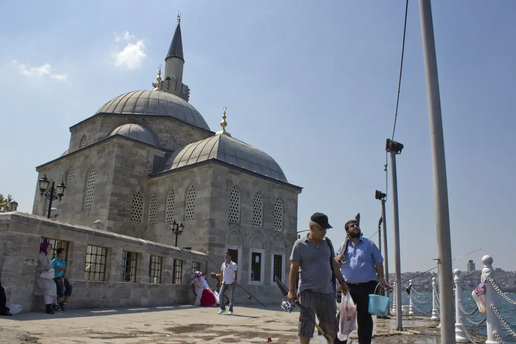 Sahil yolundan Şemsi Paşa Camii; taş duvarlar, kubbeler, minare, yürüyen insanlar ve kıyı korkulukları.