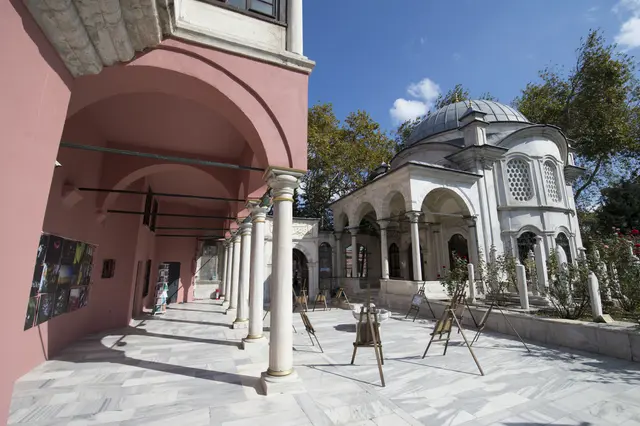 Eyüp'te Şah Sultan Külliyesi'nin avlusu; kubbeli revaklı yuvarlak türbe, mermer sütunlar, güller ve pembe mektep binası.