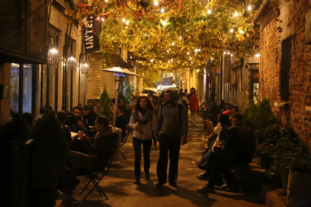 Gece Karaköy'de bir sokak; asma yapraklarının altında ışık zincirleri, masalarda oturan insanlar ve yürüyen bir çift.