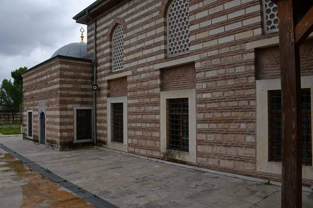Şah Sultan Camii'nin yan cephesi; almaşık duvar, petekli ve demir şebekeli pencereler, kubbeli türbe ve ıslak taş zemin.