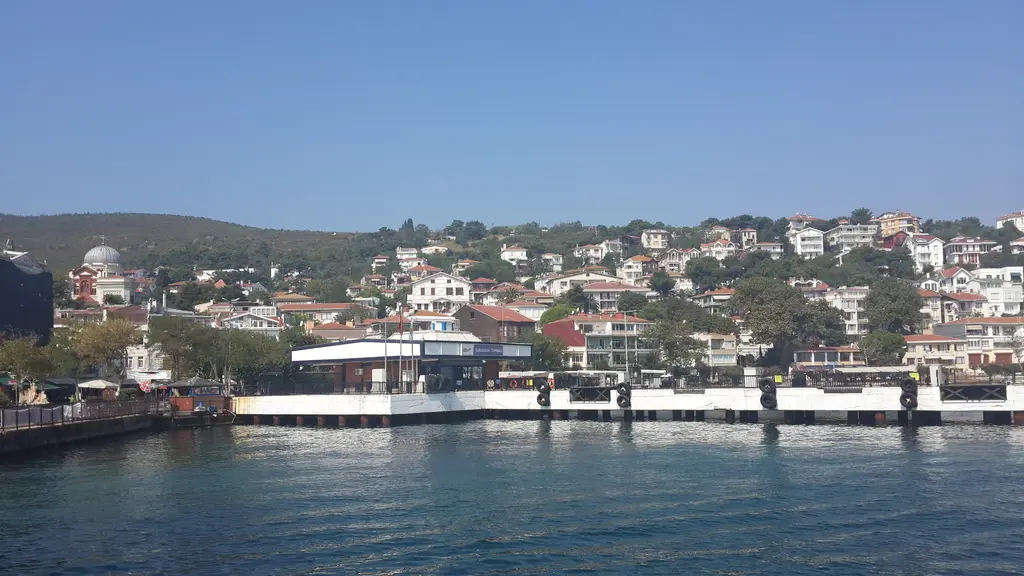 Burgazada'da yamaç; çatıların ve ağaçların arasından görülen deniz, karşı kıyıda İstanbul siluetleri.