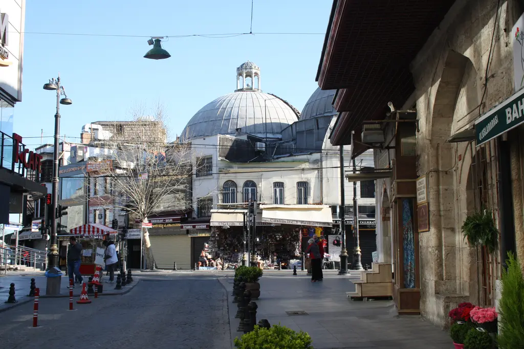 Vezirhan Caddesi'nden Çemberlitaş Hamamı; ortada kurşun kubbe, iki yanda dükkânlar, yolda bariyerler ve tente gölgesinde yürüyen insanlar.
