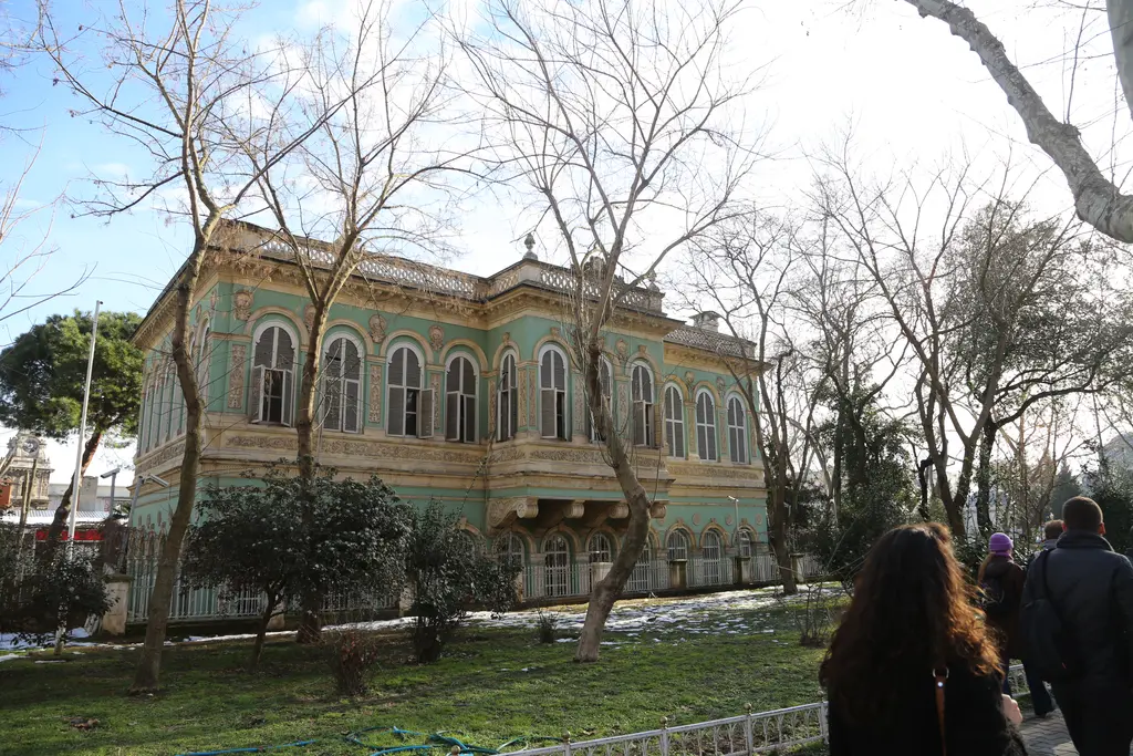 Kışın Tophane Kasrı; yapraksız ağaçların arasında yeşil köşk, çimenlikte kar lekeleri ve yürüyen insanlar.