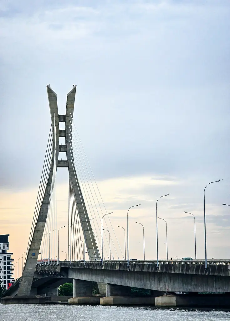 Lagos'ta Lekki-Ikoyi bağlantı köprüsünün tek pilonlu, çelik halatlı silüeti; alacakaranlıkta lagün üzerinde uzanan sokak lambalarıyla.