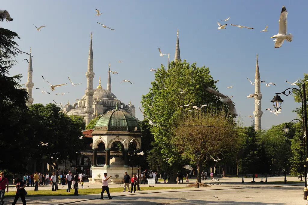 Sultanahmet Meydanı'nda Alman Çeşmesi; önde yürüyen insanlar, ağaçların arasında çeşmenin kubbesi, arkada Sultanahmet Camii ve uçan martılar.