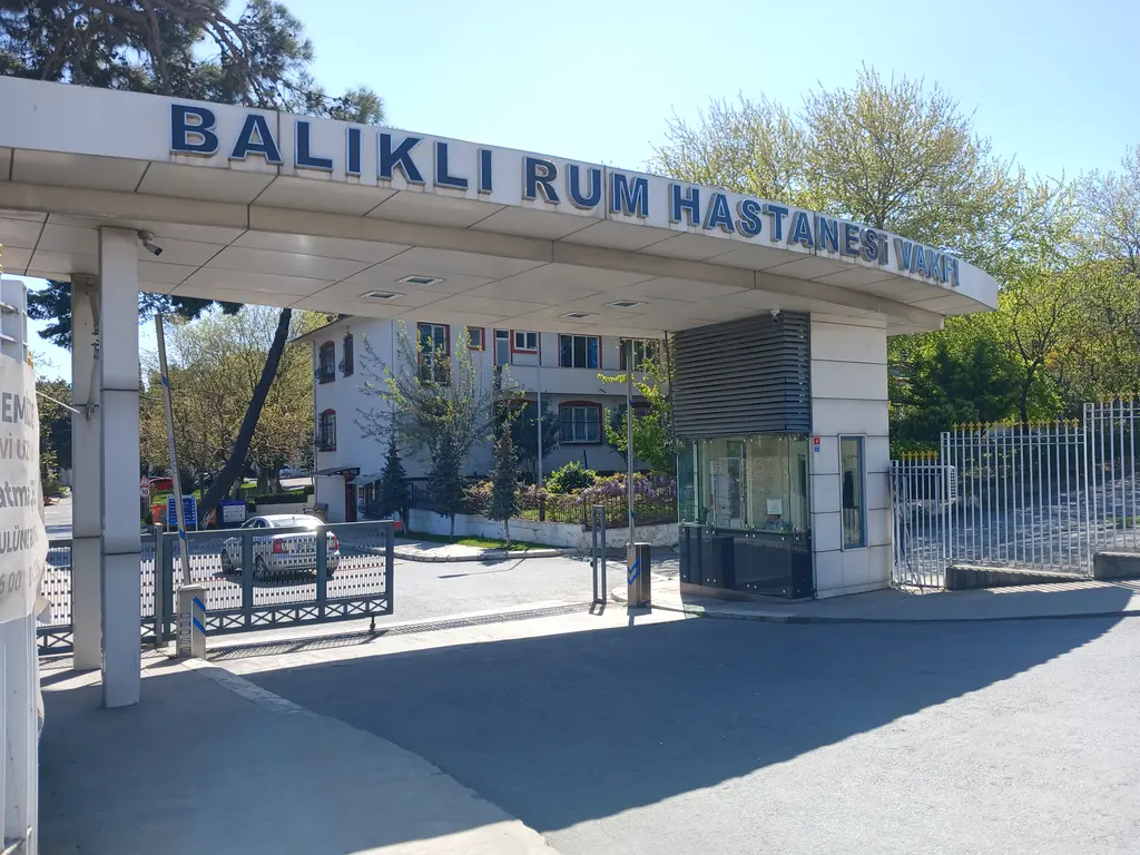 Balıklı Rum Hastanesi'nin girişi; üzerinde hastane adının yazılı olduğu beyaz saçak, güvenlik kulübesi, demir parmaklık ve ağaçlar.
