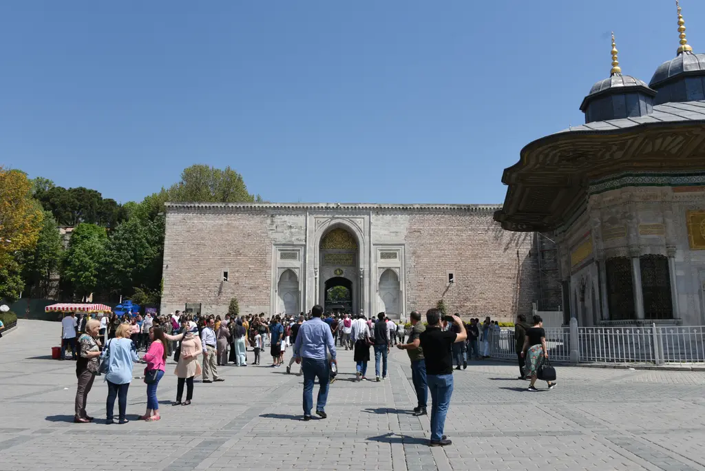 Meydan; solda Topkapı Sarayı'nın anıtsal Bab-ı Hümayun kapısı ve önündeki kalabalık, sağda çeşmenin kubbeleriyle saçağı.