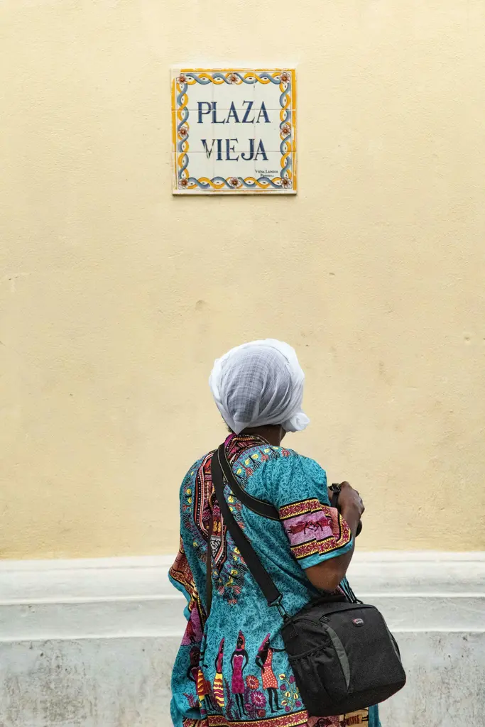 Havana'da sarı bir duvarın önünde yürüyen, desenli mavi bluz ve başörtüsü giymiş bir kadın; duvarda "Plaza Vieja" yazan seramik karo levha.