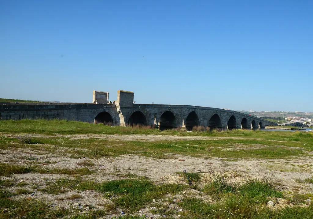Çayırdan Kanuni Sultan Süleyman Köprüsü; önde otlu düzlük, uzakta alçak kemerli köprü ve kitâbe köşkleri.