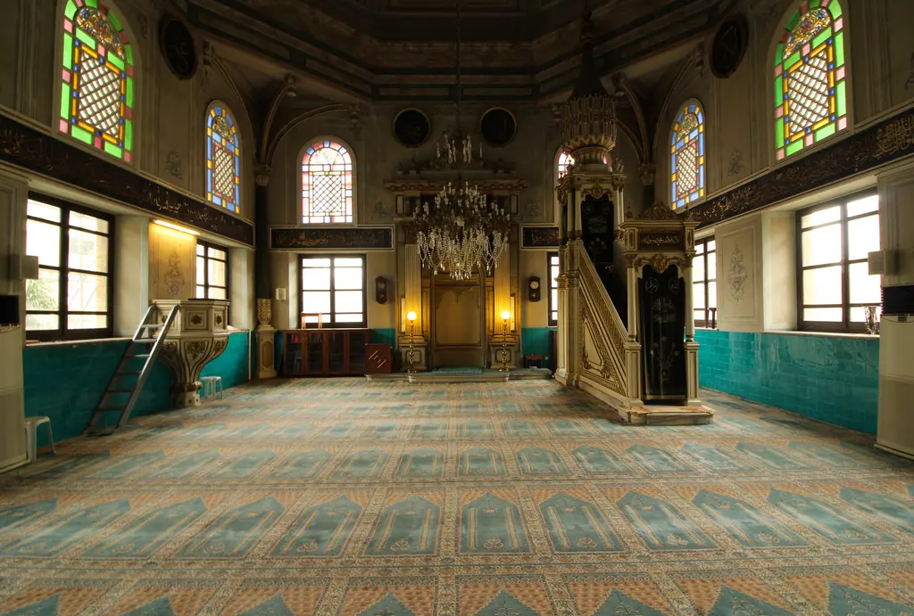 Teşvikiye Camii'nin içi; renkli vitray pencereler, yaldızlı ahşap minber ve mihrap, kristal avize, turkuaz çini bant ve desenli halı.