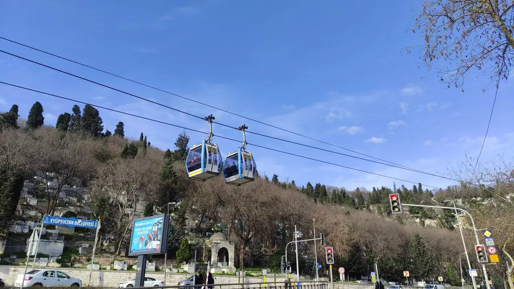 Eyüp-Piyer Loti teleferiği; mavi gökyüzünde halata asılı iki kabin, alttaki yamaçta çıplak ağaçlar ve kıyıdaki yol.