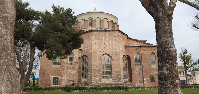 Aya İrini dışarıdan; ağaçların arasında tuğla ile taşın şerit şerit örüldüğü gövde, üstünde kurşun kaplı kubbe ve yan cephede kemerli pencere sıraları.