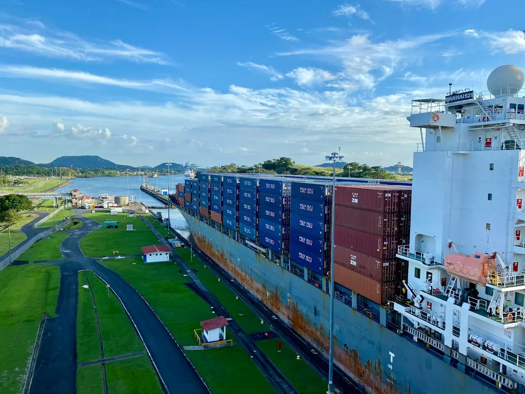 Panama Kanalı'nda bir kilit havuzundan geçen dev konteyner gemisi; rengârenk konteyner yığınları, yeşil kıyı şeridi ve açık mavi gökyüzü.