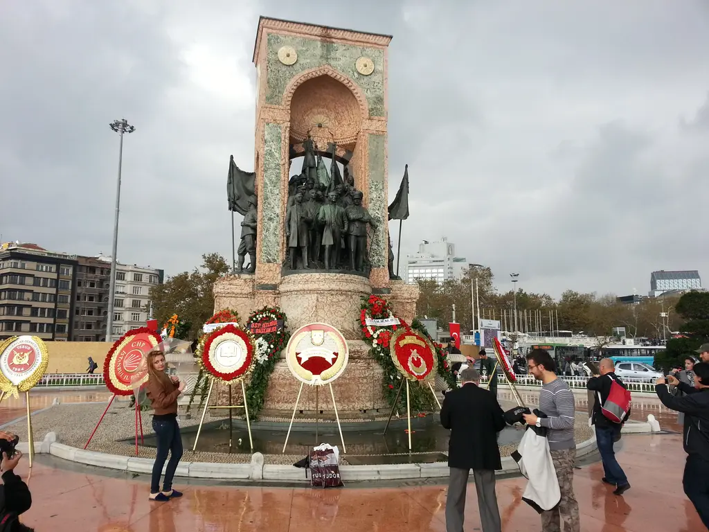 Bulutlu bir günde Cumhuriyet Anıtı; kaidesinin önünde çelenkler, anıtın fotoğrafını çeken ziyaretçiler ve ıslak zemin.
