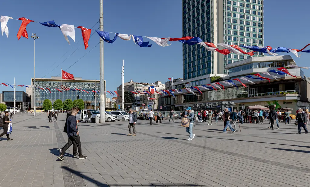 Taksim Meydanı'nda gerilmiş renkli flamalar altında yürüyen insanlar; arkada Atatürk Kültür Merkezi ve yüksek bir otel binası.