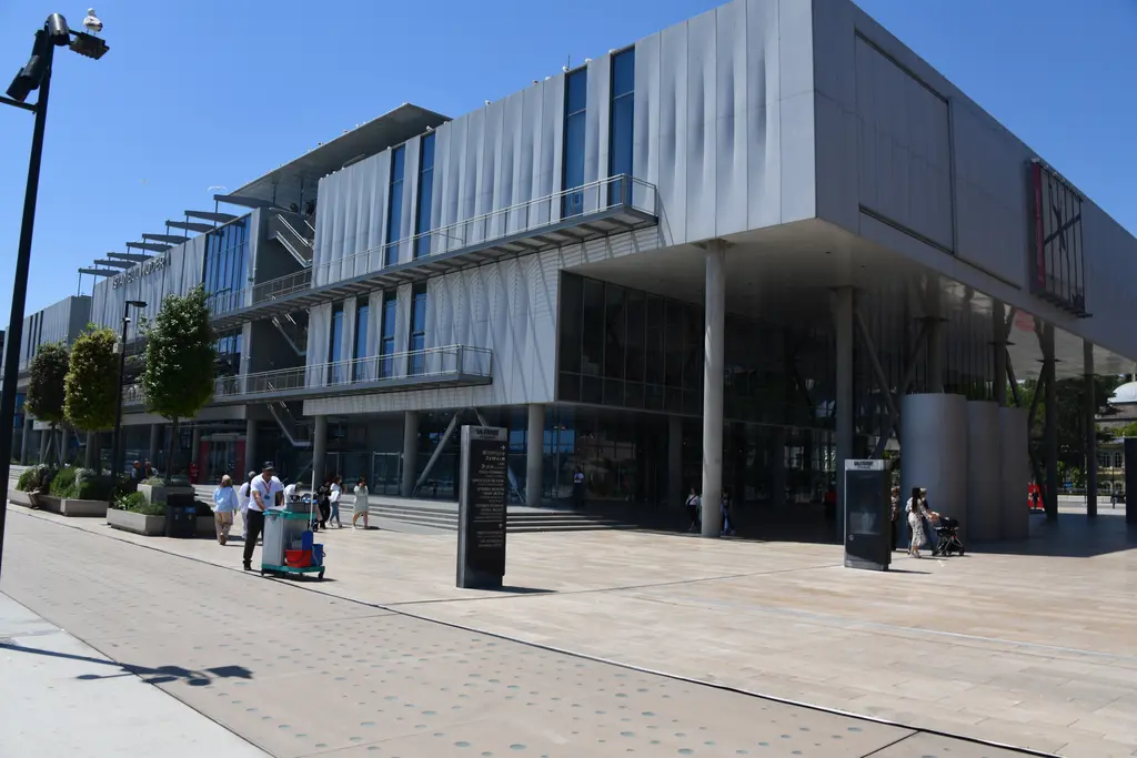 İstanbul Modern'in girişi üzerinde ileri doğru uzanan kütle; altında sütunlar, yanında dış merdiven ve kaldırımda ziyaretçiler.