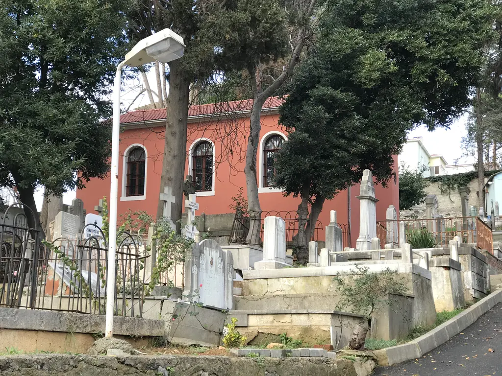 Tarabya Rum Ortodoks Mezarlığı'nda kırmızı cepheli kilise; demir parmaklık, mezar taşları ve ağaçlar.