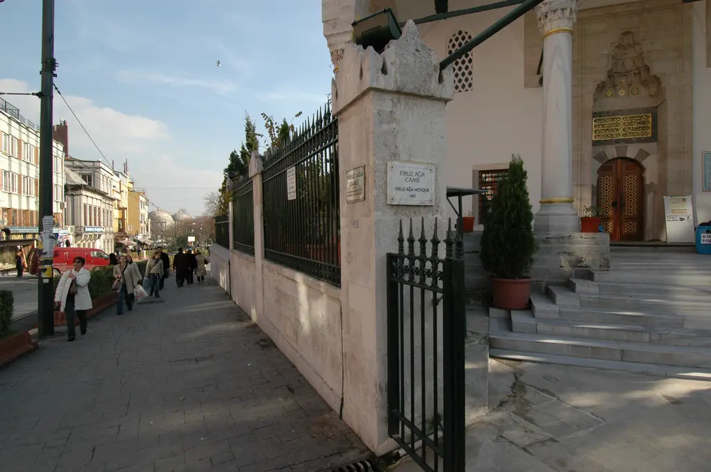 Firuz Ağa Camii'nin avlu kapısı; demir parmaklıklı duvar, taş basamaklar, saksılarda bitkiler ve kaldırımda yürüyen insanlar.