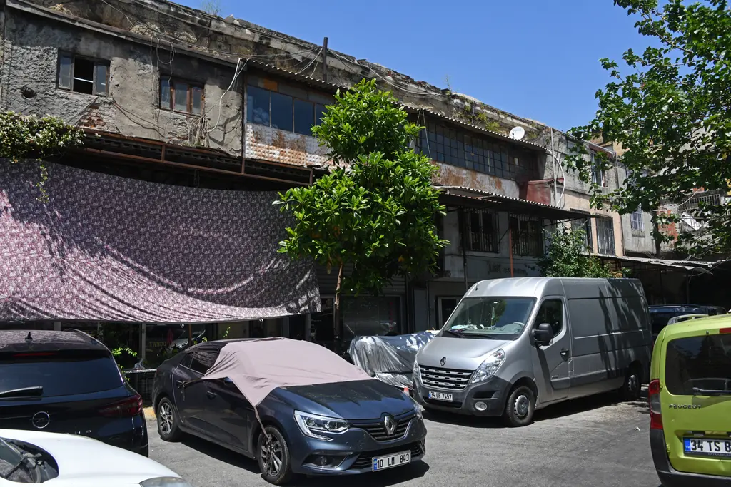 Büyük Valide Han'ın avlusu; mor desenli bir tente, ortada ağaçlar, park etmiş arabalarla minibüs ve arkada sıvası dökülmüş iki katlı han kanadı.
